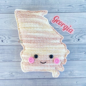 Puede incluir: Un juguete de peluche de crochet con forma del estado de Georgia con una cara sonriente y mejillas rosadas. El juguete está sobre un fondo de madera clara con una etiqueta roja y blanca a rayas que dice "Georgia".