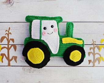 May include: A green and yellow crocheted tractor toy with a white face and black eyes. The tractor is sitting on a white wooden surface with brown paper cutouts of corn stalks behind it.