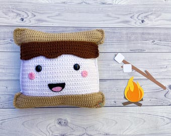 May include: A crocheted plush pillow shaped like a marshmallow with a brown chocolate topping. The pillow has a smiling face with pink cheeks and a red mouth. The pillow is on a white wooden background with a paper campfire and marshmallows.
