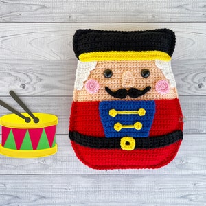 May include: A crocheted nutcracker doll wearing a red and blue uniform, a black hat, and a yellow belt. The doll is holding a small yellow drum with red and green accents.