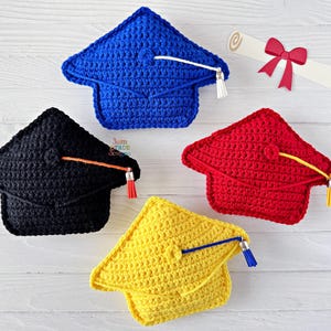 May include: Four crocheted graduation caps in blue, black, red, and yellow, each with a tassel. A rolled diploma with a red ribbon is also present. The caps are handmade and likely for a graduation celebration.