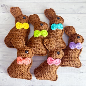 May include: Six crocheted brown bunny rabbits with colorful bows. The bows are yellow, green, blue, purple, pink, and pink.