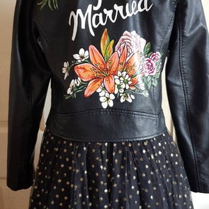 May include: A black leather jacket with a floral design and the words "Just Married" painted on the back. The jacket is paired with a black tulle skirt with gold polka dots.