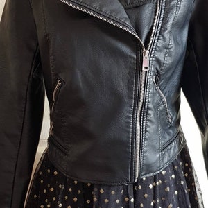 May include: Black faux leather moto jacket with a zipper closure and silver hardware. The jacket is cropped and has a collar. The jacket is worn over a black tulle skirt with gold polka dots.