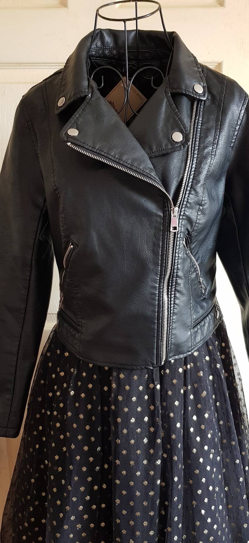 May include: Black faux leather moto jacket with a zipper closure and silver hardware. The jacket is cropped and has a collar. The jacket is worn over a black tulle skirt with gold polka dots.