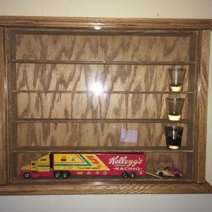 May include: A wooden display case with multiple shelves, holding toy race cars and shot glasses. The case features a Kellogg's Racing semi-truck toy. The case is made of oak wood and has a glass front.