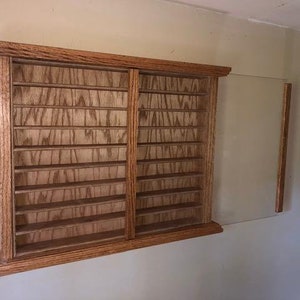 May include: A wooden display case with multiple shelves and a sliding glass door. The case is made of light brown wood and is mounted on a wall. The shelves are designed to hold small items.
