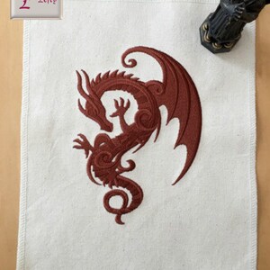 May include: A white fabric square with a red embroidered dragon design. The dragon is curled up with its wings spread out. The fabric has a slightly textured surface.