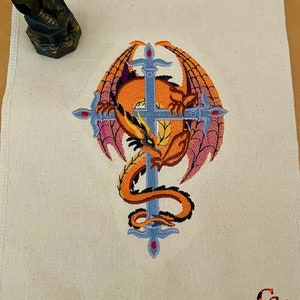 May include: A white fabric coaster with an embroidered design of an orange dragon wrapped around a silver sword with a red jewel at the tip. The dragon's wings are purple and the background is white. The coaster has the initials "C&L" embroidered in red in the bottom right corner.