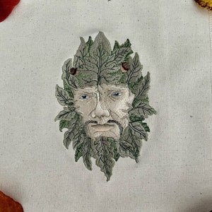 May include: A white fabric square with a green embroidered image of a man's face with leaves and acorns. The fabric has a subtle texture and is surrounded by fall leaves.