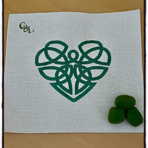 May include: A white fabric coaster with a green Celtic knot heart design and the initials "C&L" in gold thread. Three green felt balls are on the wooden surface below the coaster.