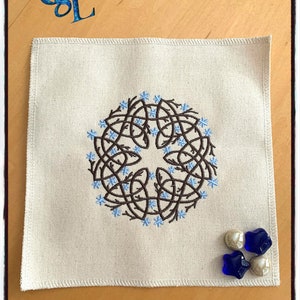 May include: A white linen square with a brown embroidered Celtic knot design. The knot is surrounded by blue flower-like shapes. There are three blue and white glass beads on the linen.
