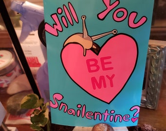 May include: A blue and pink card with the text "Will you be my Snailentine?" and a cartoon snail with a pink heart. A real snail is sitting on a wooden surface.