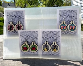 May include: A display of handmade earrings featuring insect designs. Each earring set is displayed on a playing card with a blue and white pattern. The earrings are in small embroidery hoops with images of an ant, a ladybug, a butterfly, and a firefly.