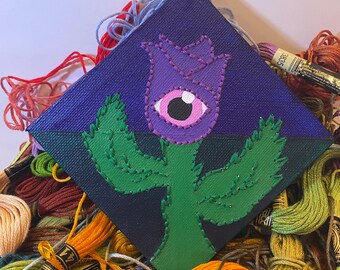May include: A hand-stitched, diamond-shaped canvas art piece featuring a purple flower with an eye in the center. The flower is on a green stem, set against a dark blue background. The artwork is surrounded by colorful embroidery floss.