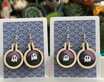 May include: A pair of handmade earrings featuring a small, embroidered ghost design within a wooden embroidery hoop. The background fabric is a dark purple, and the earrings hang from silver-colored hooks. The earrings are displayed on a patterned card.