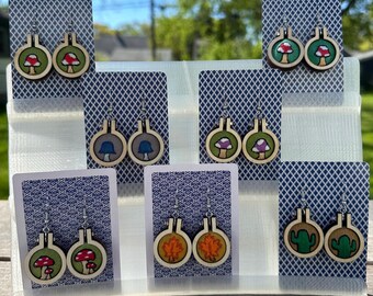 May include: A display of handmade wooden earrings with various designs. The earrings feature mushroom, leaf, and cactus motifs, each set within a circular frame. The color palette includes green, red, brown, and blue, with silver-tone earring hooks.