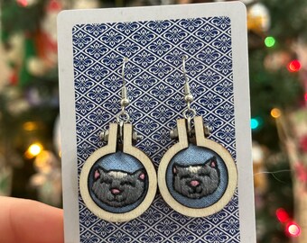 May include: A pair of handmade earrings featuring embroidered cat faces within small wooden embroidery hoops. The cats are gray with pink noses and are set against a light blue background. The earrings hang from silver-colored hooks.