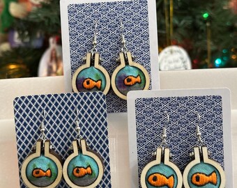 May include: Three pairs of handmade earrings featuring a wooden embroidery hoop design. Each earring showcases a small, embroidered orange fish inside a blue and teal bubble. The earrings are displayed on patterned cards.