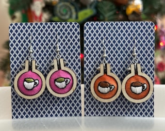 May include: Embroidered coffee cup earrings. One pair features pink fabric, the other orange. The coffee cup design is stitched in white and green thread, set in a wooden embroidery hoop. The earrings hang from silver-colored hooks.