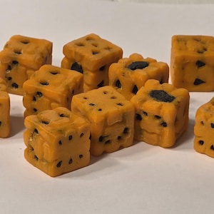 May include: A collection of small, orange, cube-shaped objects with dark spots and holes. The objects are arranged on a white surface. The cubes have a textured appearance and are evenly spaced.