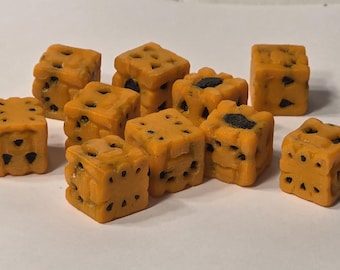 May include: A collection of small, orange, cube-shaped objects with dark spots and holes. The objects are arranged on a white surface. The cubes have a textured appearance and are evenly spaced.