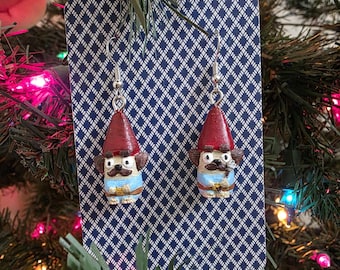 May include: A pair of handmade gnome earrings. Each earring features a gnome with a red hat, a brown mustache, and a blue shirt. The earrings are displayed on a blue and white patterned card, with silver-colored ear wires.