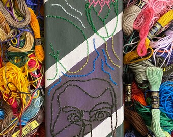 May include: A rectangular canvas with a stitched design featuring a face and abstract elements. The canvas has a purple background with white and green diagonal stripes. The artwork is surrounded by colorful embroidery floss.