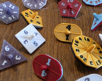 May include: A collection of colorful, geometric game pieces with numbers. The pieces are in various shapes, including squares, triangles, and ovals, and are painted in red, yellow, blue, purple, and white. Each piece has a central spinner.