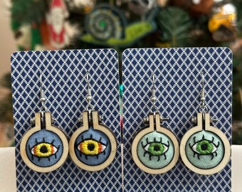May include: Embroidered eye earrings. Two pairs of earrings are displayed, each featuring a small, round embroidery hoop with a stitched eye design. One pair has yellow eyes, the other green. The hoops are set against a blue and white patterned background.