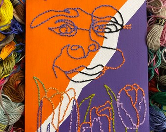 May include: An abstract embroidery art piece on canvas. The artwork features a face and tulip design, stitched with purple, black, orange, and green thread. The background is divided diagonally with orange and purple sections, with a white stripe.