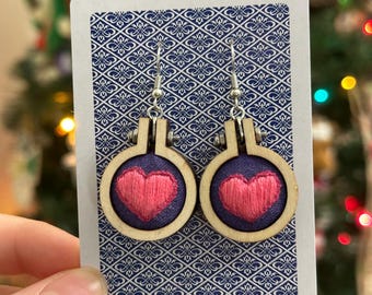 May include: A pair of handmade earrings featuring small embroidery hoops. Each hoop has a navy blue fabric background with a pink heart design. The hoops are held by silver-colored ear wires and are displayed on a patterned card.
