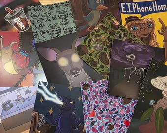 May include: A collection of colorful art prints featuring various illustrations. The prints showcase diverse themes, including a deer with glowing eyes, a dinosaur breathing lightning, and an E.T. Phone Home design. A sticker with a drink cup is also visible.