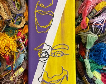 May include: A rectangular canvas art piece with a split design. One side is purple with a yellow embroidered face, the other is yellow with a black and purple embroidered face. The background is filled with colorful embroidery floss.
