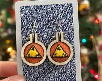 May include: A pair of handmade earrings featuring small embroidery hoops with a wooden frame. Each hoop has an orange fabric background with a yellow embroidered triangle design. The earrings are displayed on a patterned card.