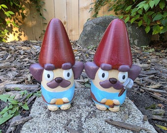 May include: Two identical garden gnome figurines featuring tall, pointed dark red hats, purple ears, and tan faces with brown mustaches. They wear light blue shirts with orange accents at the waist. One gnome holds a small gray shovel in its hand.