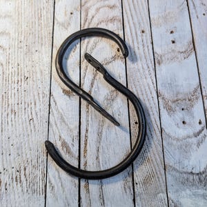 May include: A black, wrought iron S-shaped hook. The hook is made of thick metal and has a textured surface. The hook is designed to hang items and is shown against a white wooden background.