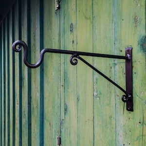 May include: A black metal wall-mounted hook with a decorative scroll design. The hook is mounted on a green wooden surface.