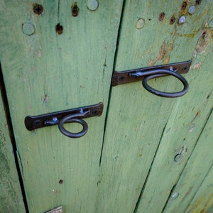 May include: Two black metal door latches with ring handles are attached to a weathered green wooden door. The door is made of horizontal planks and has several small holes and marks.