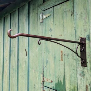May include: A wrought iron hanging bracket with a curved hook and decorative scrollwork. The bracket is mounted on a weathered, green wooden surface, suggesting outdoor use. The dark brown metal contrasts with the aged wood.