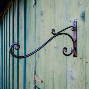 May include: A black metal hook with decorative swirls is mounted on a green wooden wall. The hook is designed to hold a hanging object.
