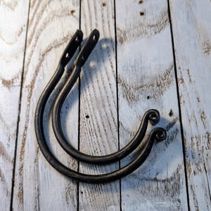 May include: Two black, curved metal hooks with a rustic, wrought iron appearance. Each hook has a looped design at the bottom and a flat top with two holes for mounting. The hooks are displayed on a weathered, white-painted wooden surface.