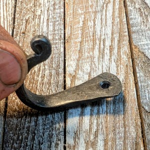 May include: Hand-forged, dark metal hook with a curved design. The hook features a decorative swirl at the top and a flat, rectangular base with a single hole. The hook is designed for hanging items and is made of durable metal.