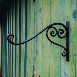 May include: Black metal wall-mounted hook with a decorative scroll design. The hook is mounted on a green wooden surface.