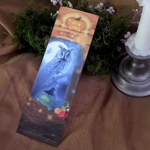 Puede incluir: Un marcapáginas con un búho azul con una rana que lleva un sombrero de bruja en primer plano. El fondo es una escena de bosque iluminado por la luna con una calabaza en la parte superior. El marcapáginas tiene el sitio web "www.artandthelittlemagic.com" impreso en la parte inferior.