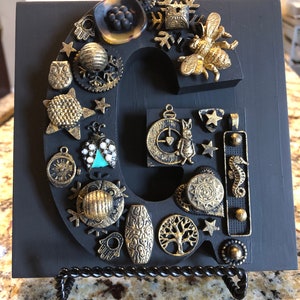 Peut inclure: Une lettre "G" en bois noir ornée de divers ornements de style antique. Une abeille dorée, des étoiles et d'autres breloques ornées sont fixées à la lettre. La lettre est présentée sur un support en métal décoratif.