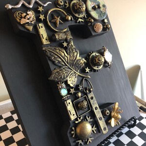 Peut inclure: Une lettre "Y" en bois noir ornée de décorations métalliques. Comprend un serpent argenté, des étoiles dorées, des engrenages, une feuille, une abeille et d'autres pièces ornées. La lettre est présentée sur une surface à carreaux noirs et blancs.