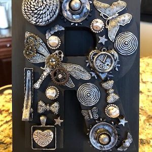 Peut inclure: Lettre "R" en bois noir décorée de divers ornements métalliques argentés et bronze. Les décorations comprennent des engrenages, des papillons, des étoiles et une libellule. La lettre est montée sur un support noir.