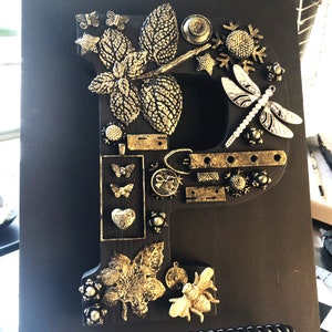 Könnte beinhalten: Schwarzer Holzbuchstabe "P" verziert mit goldfarbenen Verzierungen. Der Buchstabe ist mit Schmetterlingen, Blättern, einer Libelle und anderen dekorativen Elementen geschmückt. Der Buchstabe wird auf einer schwarzen Oberfläche angezeigt.