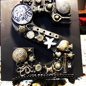 Puede incluir: Una letra "S" decorativa de madera negra adornada con varios adornos metálicos. La letra está adornada con una variedad de formas, incluyendo mariposas, corazones, un pez y una estrella de mar. La base de la letra tiene una cuerda metálica decorativa.